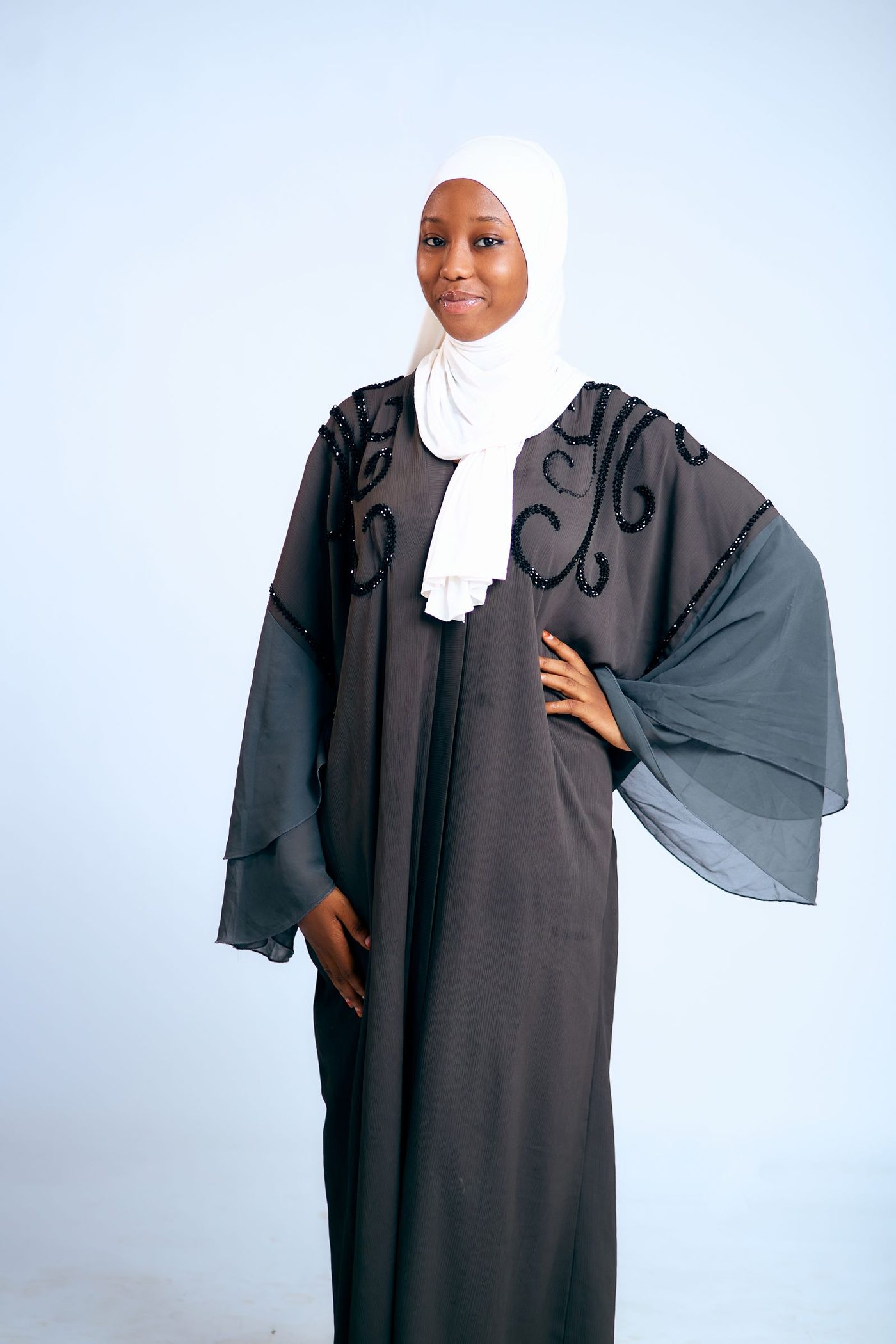 A model wearing an elegant black embellished abaya with a white hijab, studio portrait